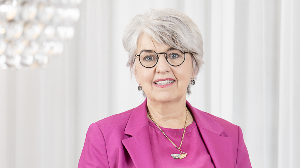 Portrait photographique de la conseillère fédérale Elisabeth Baume-Schneider. Vêtue d’une veste rose fuchsia et d’un pantalon noir, elle pose la main droite sur une table en bois et sourit à la caméra. L’arrière-plan est clair, avec des rideaux blancs et un plafonnier moderne en forme de boule.