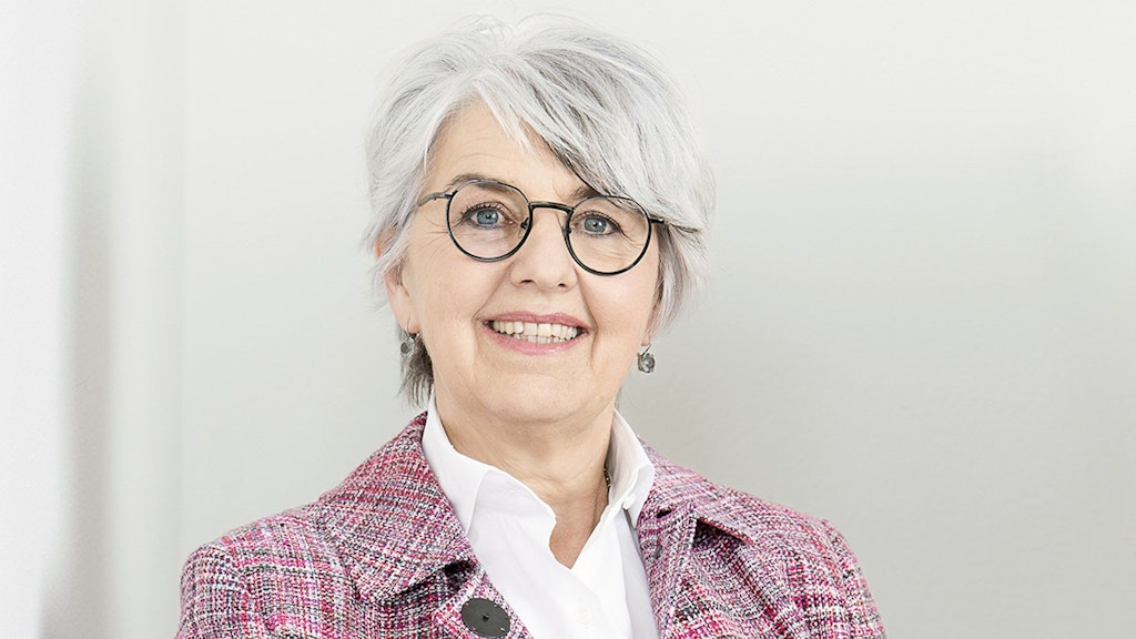 Portrait photographique de la conseillère fédérale Elisabeth Baume-Schneider. Vêtue d’un blazer rose chiné et d’un pantalon noir, elle appuie son bras sur un meuble et sourit à la caméra. L’arrière-fond est blanc.
