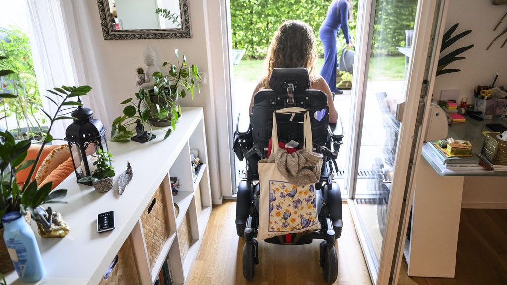 Una persona in sedia a rotelle e seduta in un appartamento luminoso davanti a una porta a vetri aperta e guarda verso il giardino, mentre un altra persona si trova sulla terrazza.