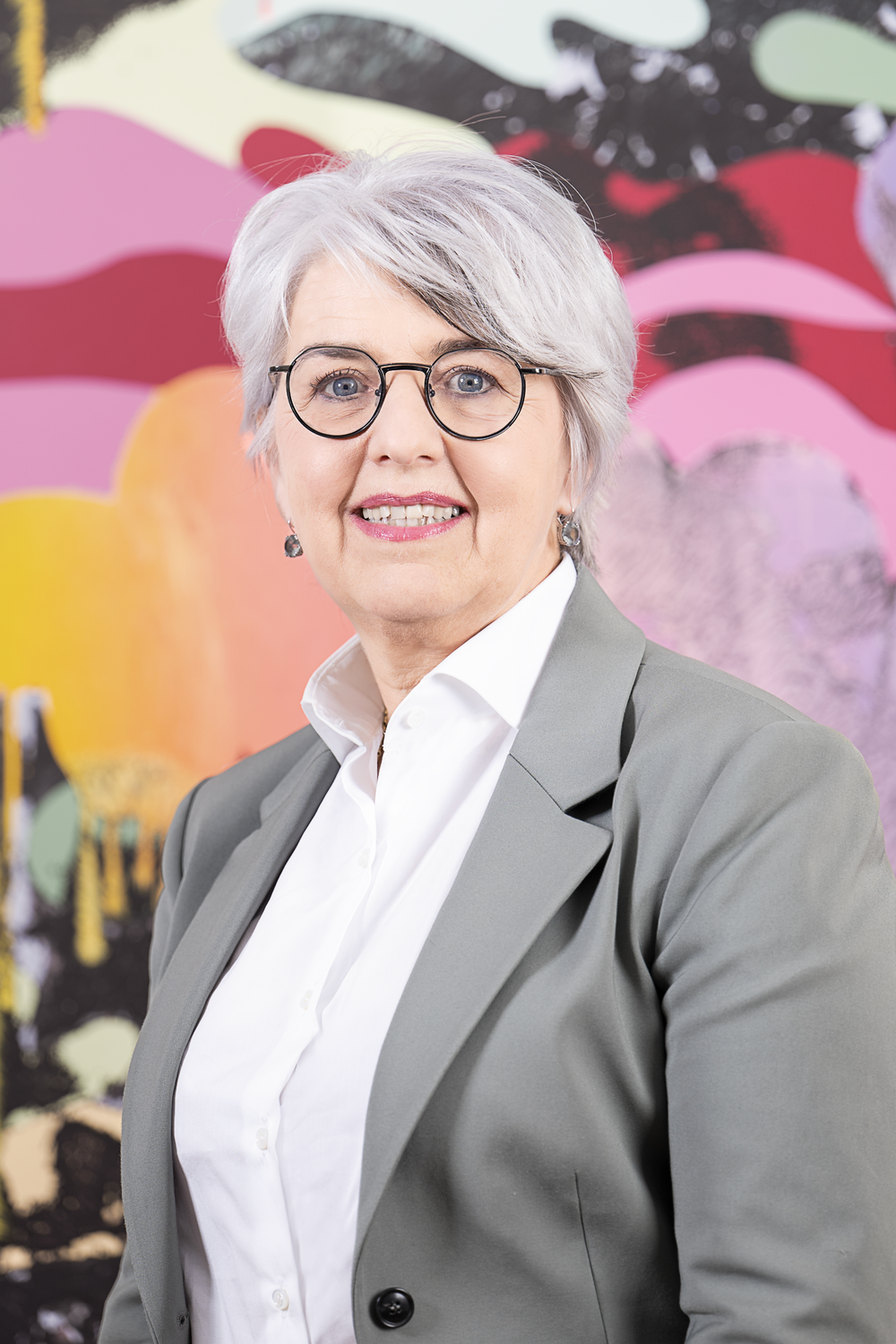 Portraitfoto von Bundesrätin Elisabeth Baume-Schneider. Sie trägt einen hellgrauen Blazer und lächelt in die Kamera. Im Hintergrund ist ein farbenfrohes Gemälde.