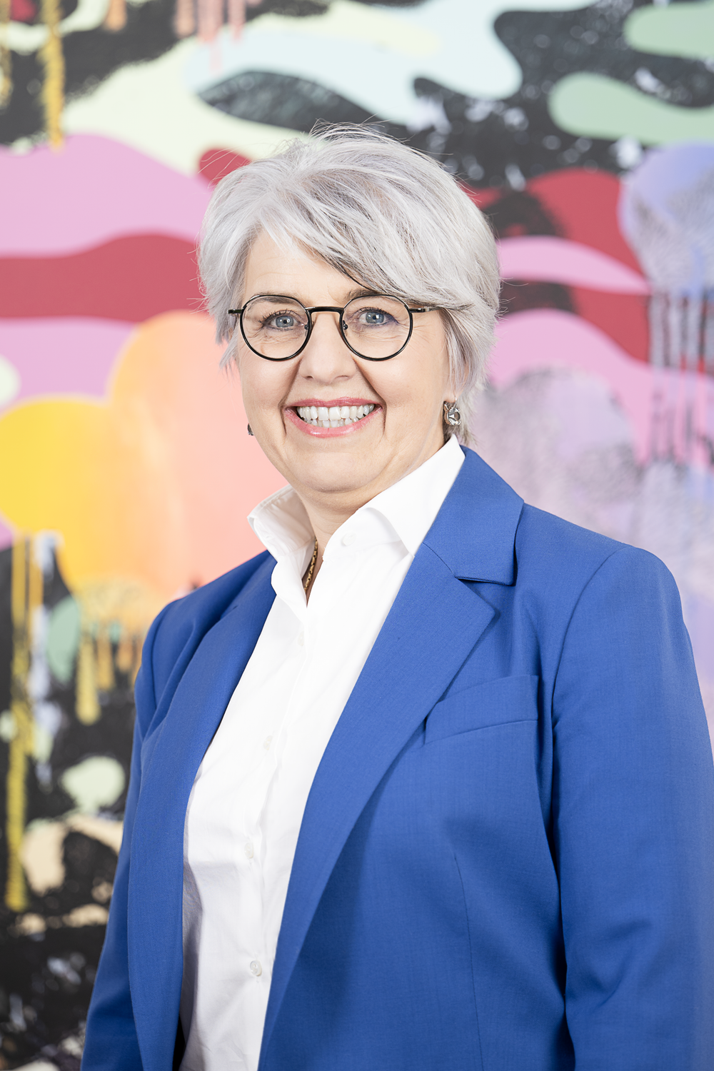 Portraitfoto von Bundesrätin Elisabeth Baume-Schneider. Sie trägt einen blauen Blazer und lächelt in die Kamera. Im Hintergrund ist ein farbenfrohes Gemälde.