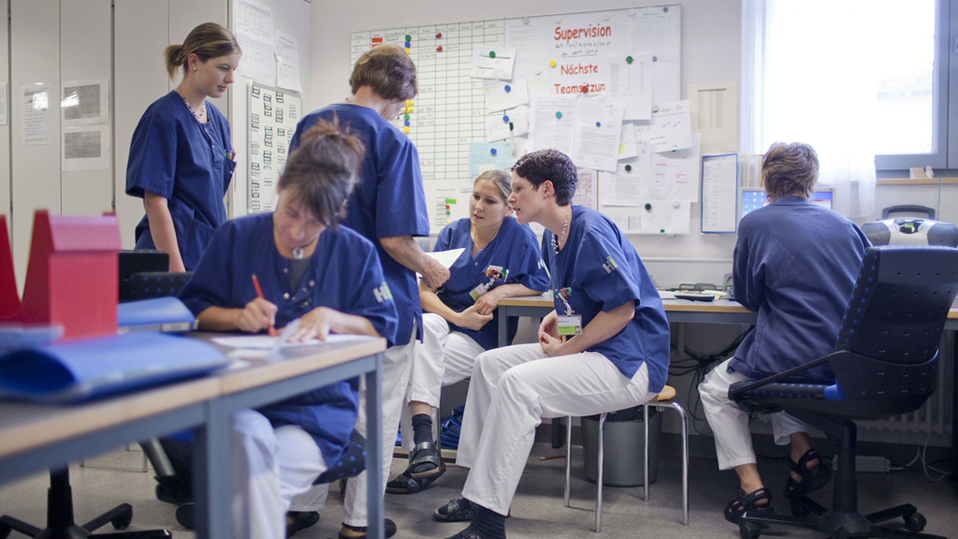 Pflegemitarbeiterinnen eines Krankenhauses halten eine Sitzung ab in einem Raum. Sie tragen alle die gleiche Arbeitskleidung.