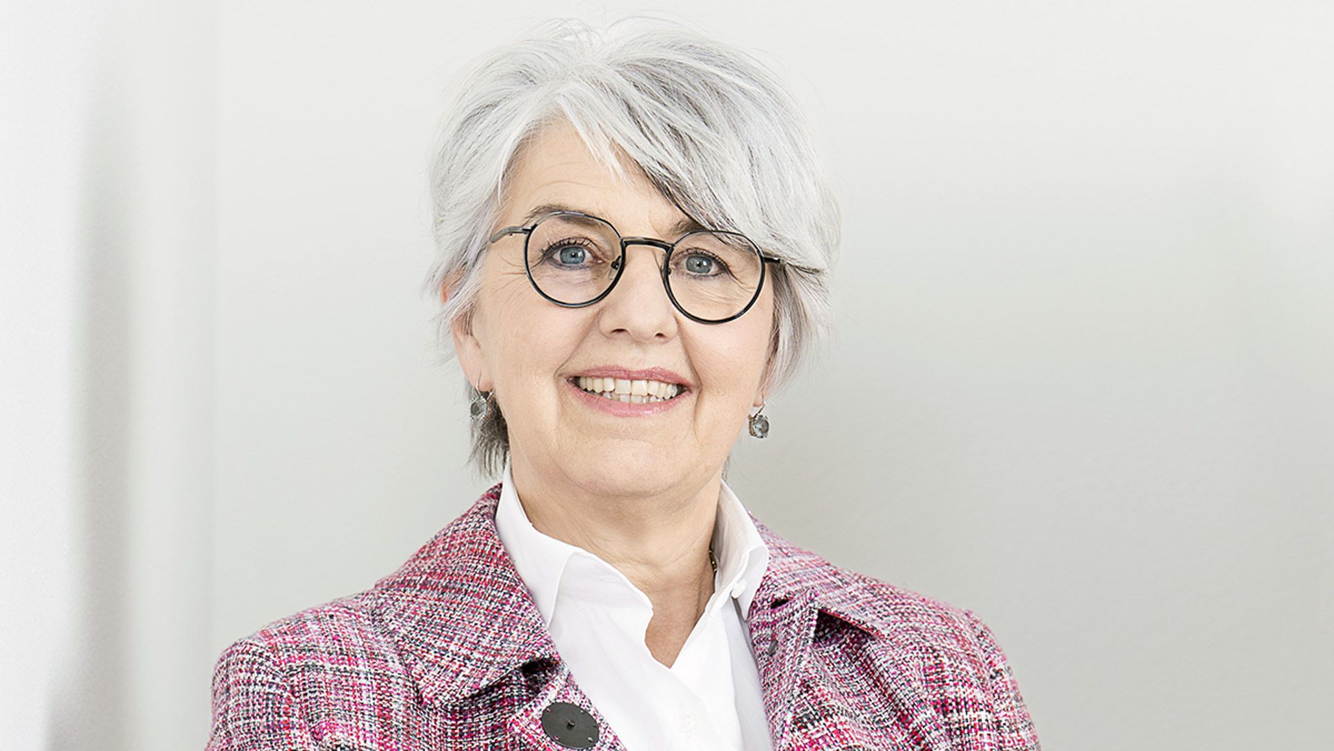 Portraitfoto von Bundesrätin Elisabeth Baume-Schneider. Sie trägt einen pinken Blazer und eine schwarze Hose. Ihren rechten Arm ist auf ein Regal gelegt. Sie lächelt in die Kamera. Der Hintergrund ist weiss.