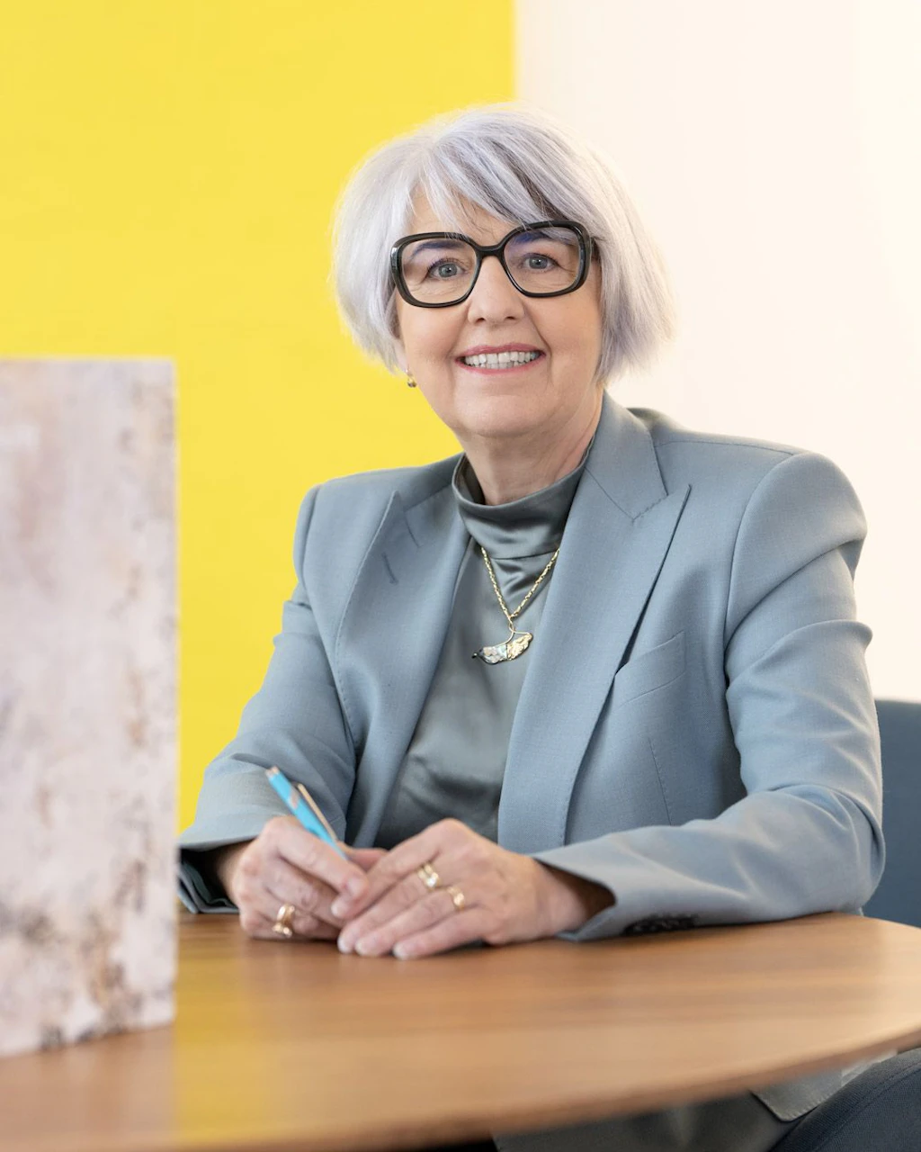 Portrait photograph of Federal Councillor Elisabeth Baume-Schneider. She is wearing a light blue blazer with black trousers. Her right hand rests on a wooden table. She is smiling into the camera. The background is well lit, with white curtains and a modern, spherical pendant lamp.