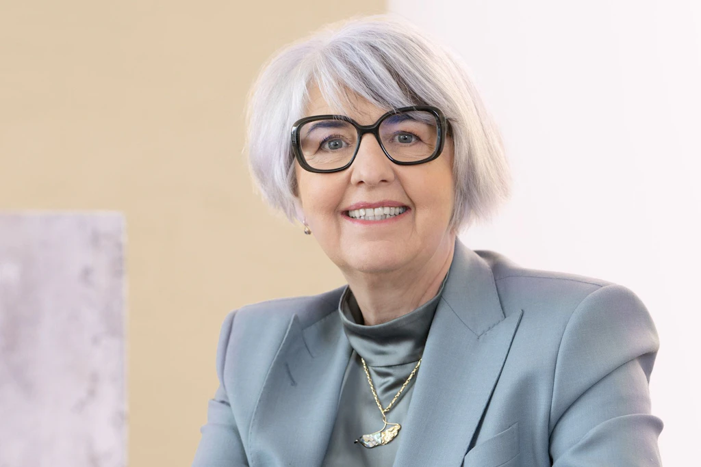 Portrait photograph of Federal Councillor Elisabeth Baume-Schneider. She is wearing a pink blazer with black trousers. Her right hand rests on a wooden table. She is smiling into the camera. The background is well lit, with white curtains and a modern, spherical pendant lamp.