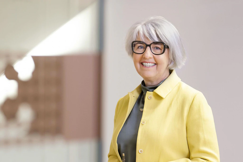 Portrait photograph of Federal Councillor Elisabeth Baume-Schneider. She is wearing a blue blazer and smiling into the camera. A colourful painting hangs in the background.