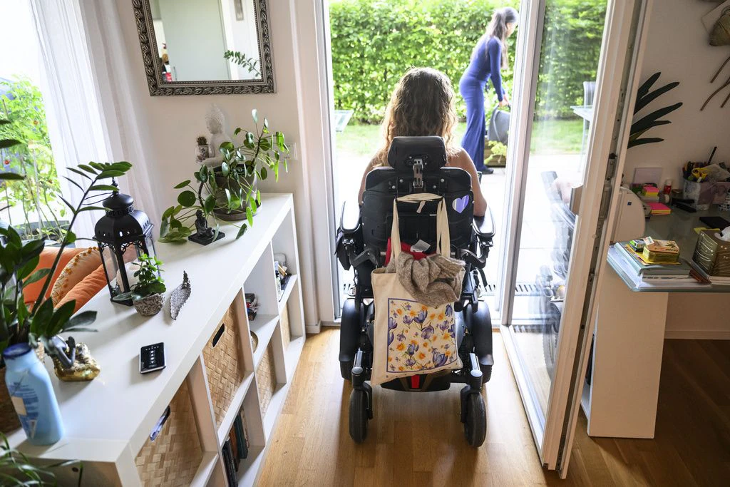 Une personne en fauteuil roulant est assise dans un appartement lumineux devant une porte vitree ouverte et regarde vers le jardin, tandis qu une autre personne se tient sur la terrasse.