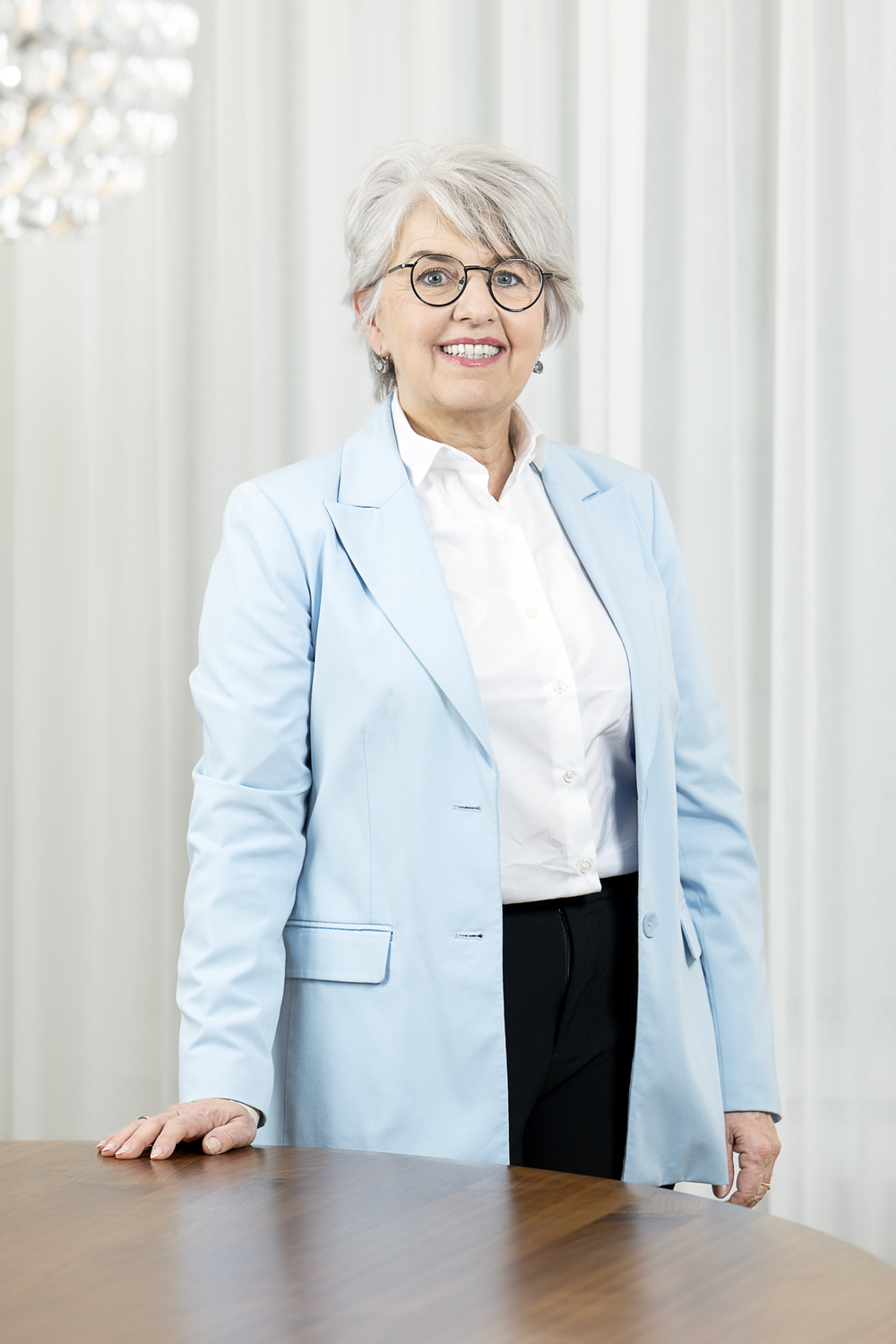 Portrait photographique de la conseillère fédérale Elisabeth Baume-Schneider. Vêtue d’un blazer bleu clair et d’un pantalon noir, elle pose la main droite sur une table en bois et sourit à la caméra. L’arrière-plan est clair, avec des rideaux blancs et un plafonnier moderne en forme de boule.