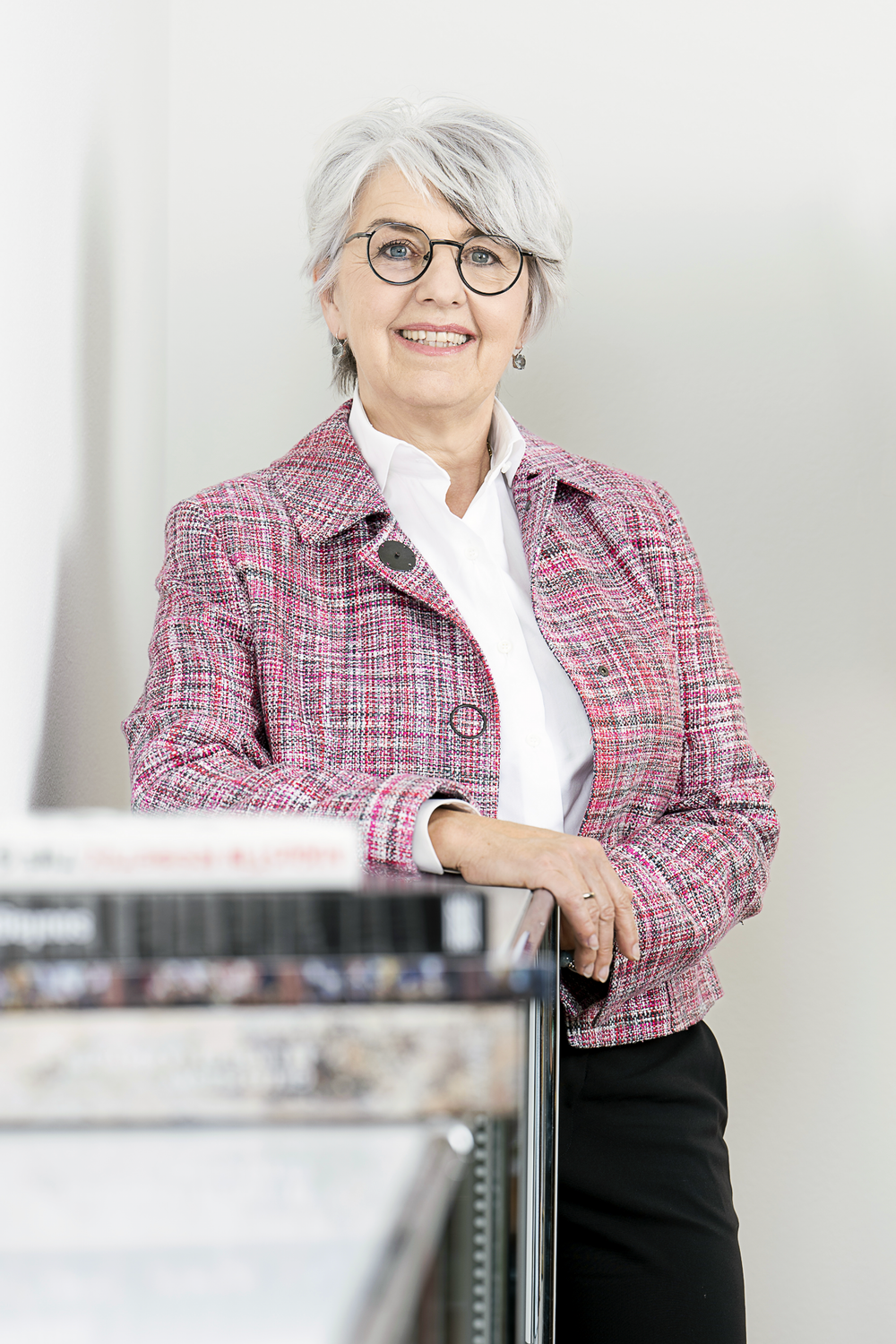 Portrait photographique de la conseillère fédérale Elisabeth Baume-Schneider. Vêtue d’un blazer rose chiné et d’un pantalon noir, elle appuie son bras sur un meuble et sourit à la caméra. L’arrière-fond est blanc.