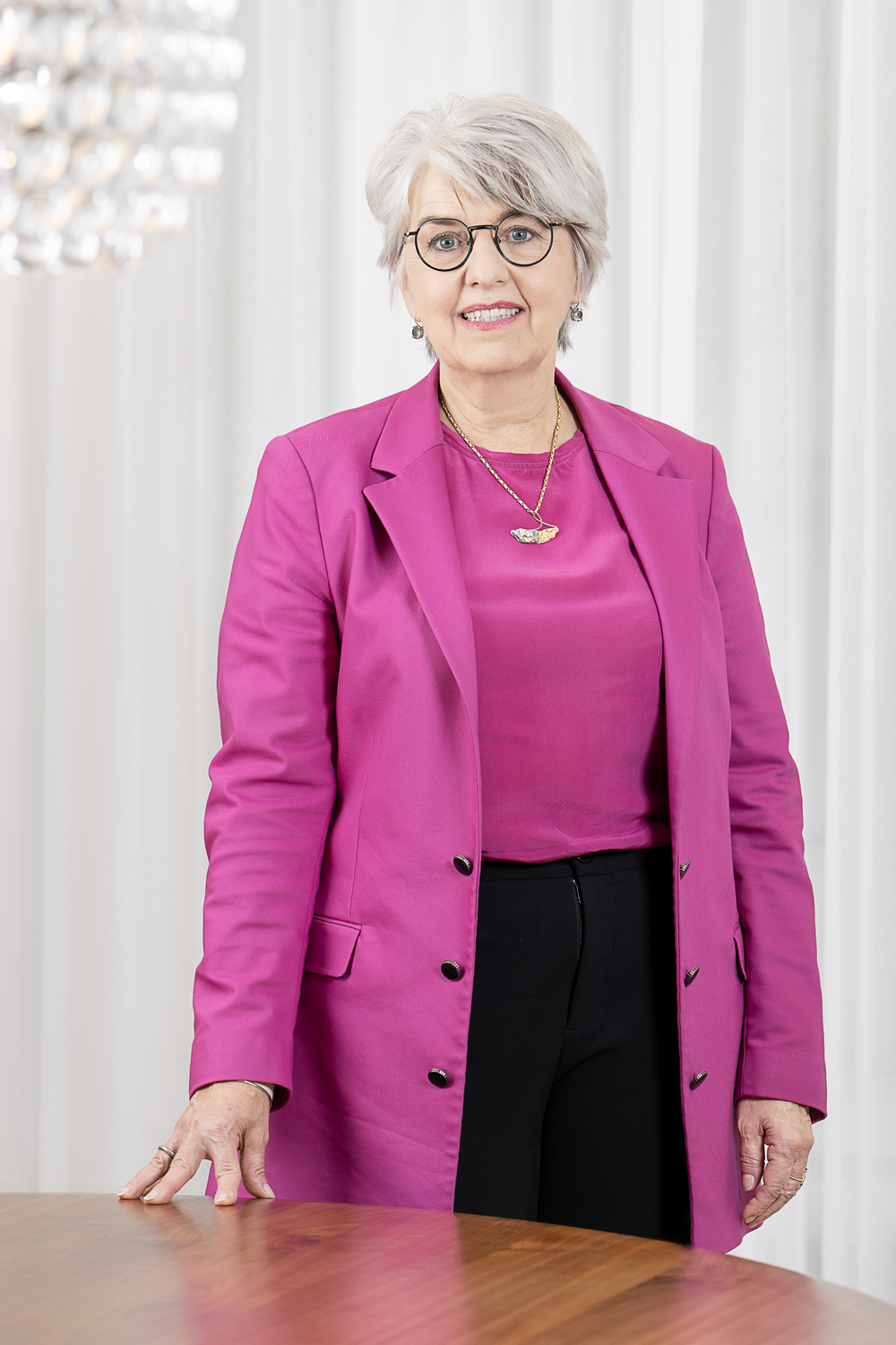 Fotoritratto della consigliera federale Elisabeth Baume-Schneider, che indossa un blazer rosa e un pantalone nero, ha la mano destra appoggiata su un tavolo di legno e sorride alla fotocamera. Sullo sfondo luminoso alle sue spalle appaiono una tenda bianca e un moderno lampadario rotondo.